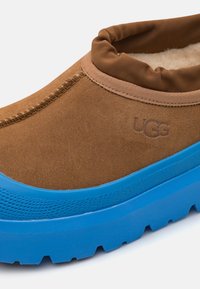 Brūns zamšādas slip-on apavi ar mīkstu, nolokāmu apkakli un zilu gumijas zoli, iekļaujot strukturālas akcentus un UGG logotipu.