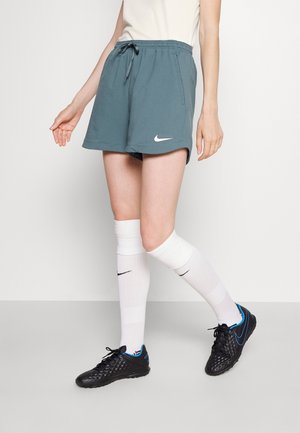 Osoba nosząca szare sportowe spodenki Nike, białe skarpetki za kolano z czarnymi logo Nike oraz czarne sportowe buty Nike z niebieskimi akcentami.