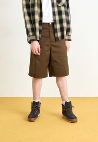 Pantaloni scurți din bumbac maro, cu o croială dreaptă, purtați cu un tricou alb și o cămașă în carouri alb-negru, asortați cu cizme din ante bleumarin.