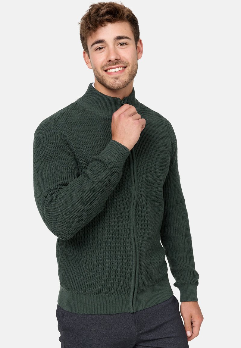 Cárdigan verde de punto con cremallera, que presenta un cuello alto, puños acanalados y un patrón texturizado. Hecho de un hilo grueso para proporcionar calidez.