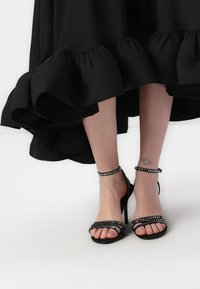 Sandales à talons hauts noires avec plusieurs sangles brillantes, des sangles pour la cheville avec des embellissements et une semelle à texture lisse.