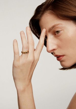 Anillo de oro con un diseño texturizado y facetado, llevado en un dedo delgado, contra un fondo neutro, con una vista lateral del rostro de una persona.