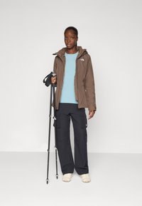 Bruine waterdichte jas met capuchon, lichtblauwe top, donkere cargobroek en beige schoenen; houdend trekkingstokken; gladde textuur.