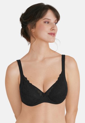 Frau mit dunklen Haaren in einem Hochsteckfrisur, die einen schwarzen Spitzen-Bügel-BH trägt und seitlich blickt, vor einem schlichten hellen Hintergrund.