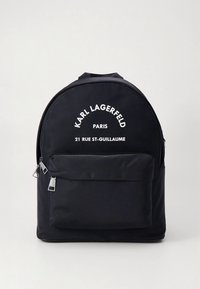 Mochila preta de nylon com formato arredondado, bolso frontal com fecho de correr e texto impresso a branco: "Karl Lagerfeld Paris 21 Rue St-Guillaume." Detalhes em ferragens prateadas.