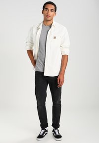 Camicia bianca a maniche lunghe sopra una t-shirt grigia, abbinata a jeans neri e sneakers nere. Da notare la tasca sul petto della camicia e il dettaglio del logo.