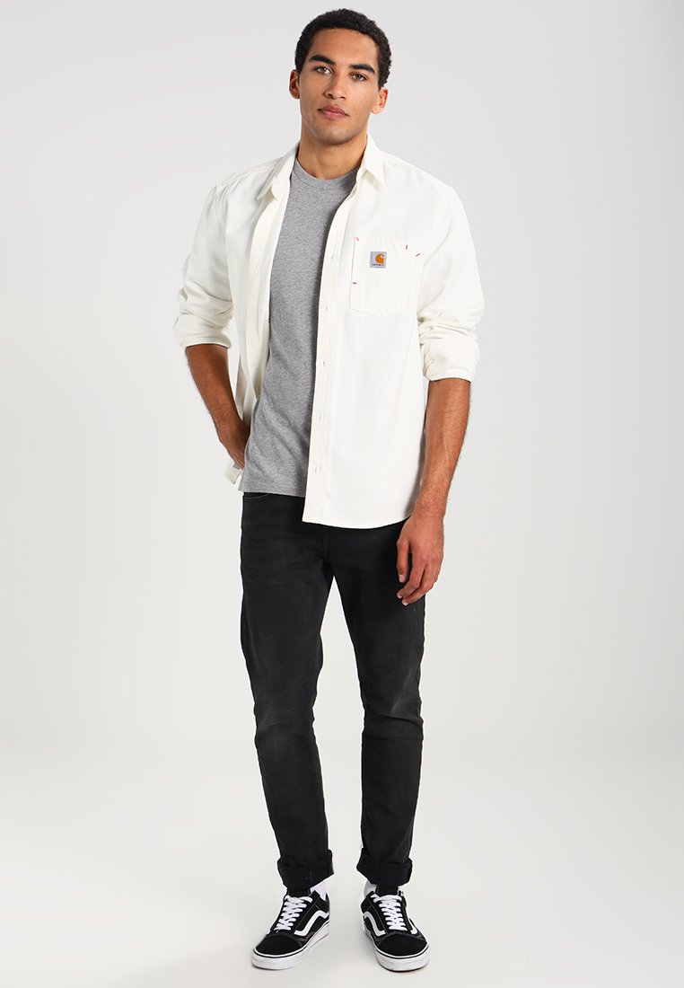 Camicia bianca a maniche lunghe sopra una t-shirt grigia, abbinata a jeans neri e sneakers nere. Da notare la tasca sul petto della camicia e il dettaglio del logo.