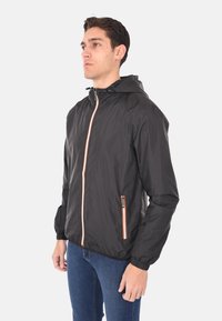 Giacca nera leggera con cappuccio, zip frontale con dettagli arancioni, polsini elasticizzati e tasche laterali per riporre gli oggetti.