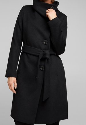 Femme portant un manteau en laine noir ceinturé avec de gros boutons et un col montant, tenant le col d'une main.