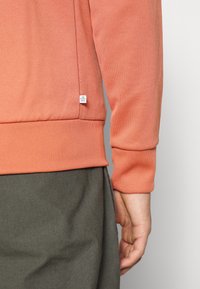 Persikofärgad sweatshirt med ribbad mudd vid ärmslut och nederkant. Mjuk textur, med en sidotagg märkt "ECO PLASTIC MADE." Bärs över grå byxor.