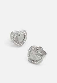 Silver heart-shaped earrings with a shiny surface, surrounded by clear rhinestones, featuring the engraved logo "GUESS" on the inner side.