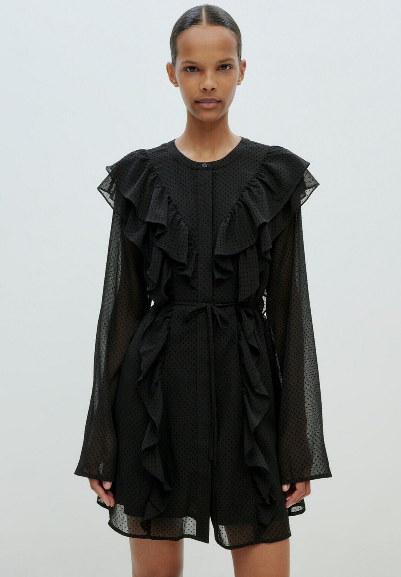 Schwarzes Kleid aus transparenter, gepunkteter Stoff, mit Rüschendetails entlang der Vorderseite und Ärmel, tailliert mit einem Bindeband und einem runden Halsausschnitt.