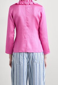 Blusa rosa con texture, colletto a volant e maniche lunghe, abbinata a pantaloni a gamba larga a strisce blu e bianche, con dettagli di bottoni sul retro.