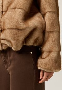 Chaqueta de pelo sintético marrón con una apariencia texturada y suave, con mangas anchas y un dobladillo redondeado, combinada con pantalones marrones oscuros.