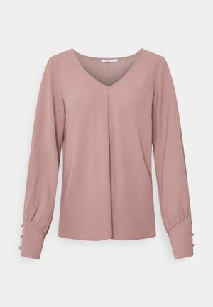 Blouse rose poudre à manches longues avec col en V, légère pince frontale et détails de boutons sur larges poignets, présentée sur fond blanc.