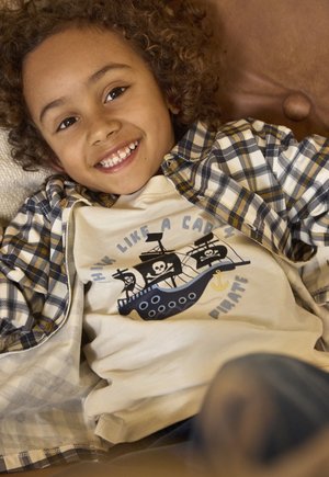 Camiseta color crema con un gráfico de un barco pirata con calaveras y un ancla. Combinada con una camisa de franela a cuadros azul y amarilla.