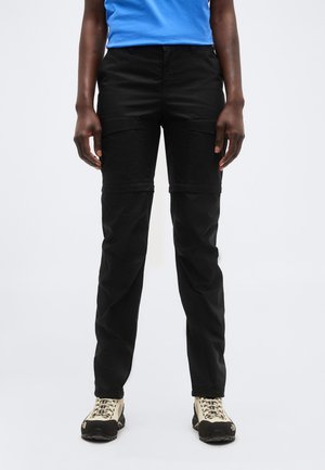 Persoană purtând pantaloni cargo negri convertibili și pantofi de drumeție bej, stând pe un fundal simplu și deschis la culoare.