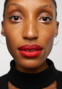 Matte rød leppestift på fyldige lepper, varm hudtone, nærbilde av munnen; subtil øyemakeup; svart høyhalset genser fremhever ansiktstrekkene.