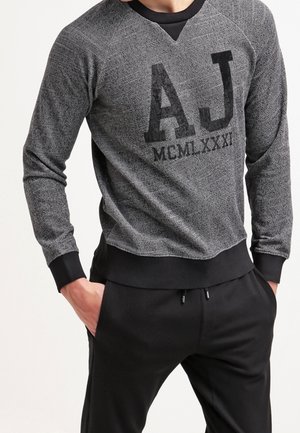 Sweatshirt noir et gris avec un motif texturé, présentant un grand logo "AJ" et le texte "MCMLXXXI". Manches et ourlet côtelés, col rond.