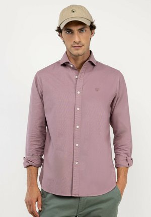 Hombre con gorra de béisbol marrón claro, camisa malva abotonada con las mangas remangadas y pantalones verdes, de pie sobre un fondo blanco liso.