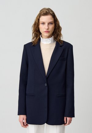 Blazer navy con hombros estructurados, cierre de un botón y dos bolsillos frontales. Se lleva sobre un suéter de color claro con un cuello con_volantes.