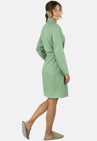 Grüner Bademantel mit Waffelstruktur und langen Ärmeln, getragen über hellgrauen Hausschuhen, aus seitlicher Perspektive präsentiert.