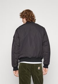 Giacca bomber nera con colletto e polsini a coste, caratterizzata da una superficie liscia e una silhouette aderente, indossata sopra pantaloni in velluto a coste verde oliva.