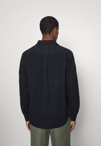 Uomo con una maglietta nera a maniche lunghe e pantaloni verde oliva, in piedi rivolto verso un muro bianco con le spalle alla macchina fotografica.