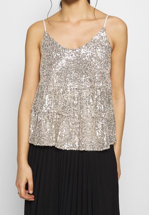 Kobieta w srebrnym, cekinowym topie bez rękawów z cienkimi ramiączkami i warstwowym dołem peplum, w połączeniu z czarną plisowaną spódnicą.