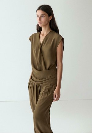 Giovane donna con lunghi capelli scuri che indossa un top verde oliva senza maniche drappeggiato e pantaloni larghi abbinati, in piedi davanti a uno sfondo bianco semplice.