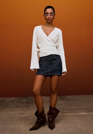 Woman wearing clear glasses, white wrap blouse with long sleeves, dark denim mini skirt, and brown slouchy boots standing on a concrete floor.