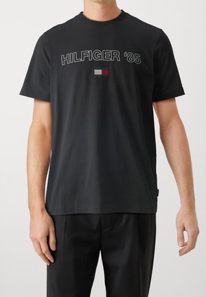 Sort bomulds T-shirt med korteærmer, der har teksten "HILFIGER '85" og et rektangulært logo badge i rød, hvid og marineblå.