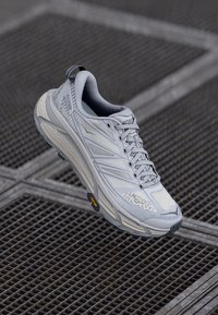 Hellgrauer Sportschuh mit Obermaterial aus Mesh und gepolstertem Schaftrand, ausgestattet mit reflektierenden Streifen und Vibram-Laufsohle, die für Dämpfung und Unterstützung konzipiert ist.