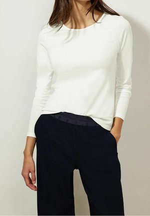 Žena má na sobě jednoduché bílé tričko s dlouhým rukávem zastrčené do vysokých černých kalhot a stojí s jednou rukou v kapse.