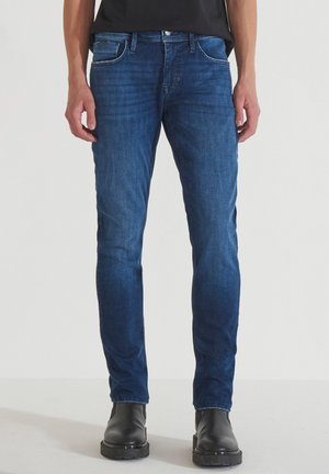 Jeans en denim bleu foncé avec une coupe ajustée, une conception à cinq poches, des coutures blanches contrastantes et des détails usés. Assortis avec des bottines noires.