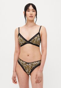 Donna che indossa un reggiseno e pantaloni intimi leopardati con pizzo nero, in piedi contro uno sfondo bianco uniforme.