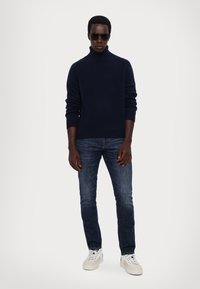 Marinefarbener Rollkragenpullover, dunkelblaue Jeans und weiße Sneaker. Der Pullover hat gerippte Bündchen; eine Sonnenbrille vervollständigt den Look.