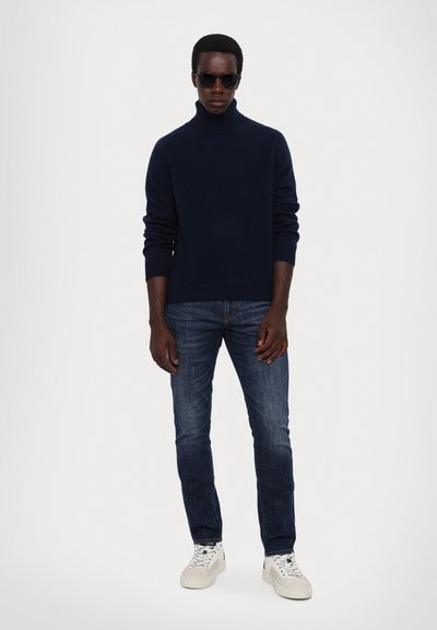 Navy turtleneck sweater, dark denim jeans, and white sneakers. The sweater features ribbed cuffs; sunglasses complete the look.