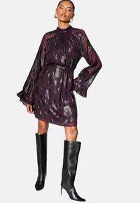 Durchsichtiges lilafarbenes Kleid mit metallic floralem Muster, hohem Kragen und langen Ärmeln, tailliert und kombiniert mit schwarzen knielangen Stiletto-Stiefeln.
