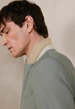 Young man with short dark hair wearing a light gray jacket with a beige collar, facing left with eyes closed against a plain beige background.