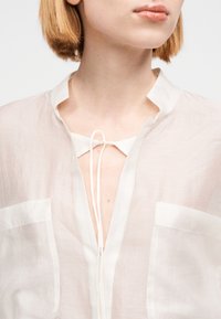 Blusa ligera y transparente en beige pálido, con escote en V con lazo en el cuello, mangas cortas y dos bolsillos delanteros. Textura suave.