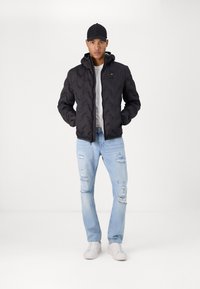 Giubbotto piumino nero con cappuccio, caratterizzato da un motivo a chevron. Indossato sopra una camicia bianca, abbinato a jeans chiari strappati e sneakers bianche.