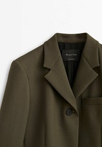 Blazer vert olive foncé sur mesure avec revers crantés, fermeture à un bouton noir, poche poitrine et doublure intérieure noire.