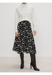 Mujer vistiendo un suéter de punto blanco metido dentro de una falda midi plisada negra con manchas abstractas blancas y botas de tacón altas negras hasta la rodilla.