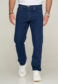 Man som bär mörkblå jeans, vita sneakers, vit skjorta, silverfärgad armbandsklocka och svart pärlarmband står på ett reflekterande golv.