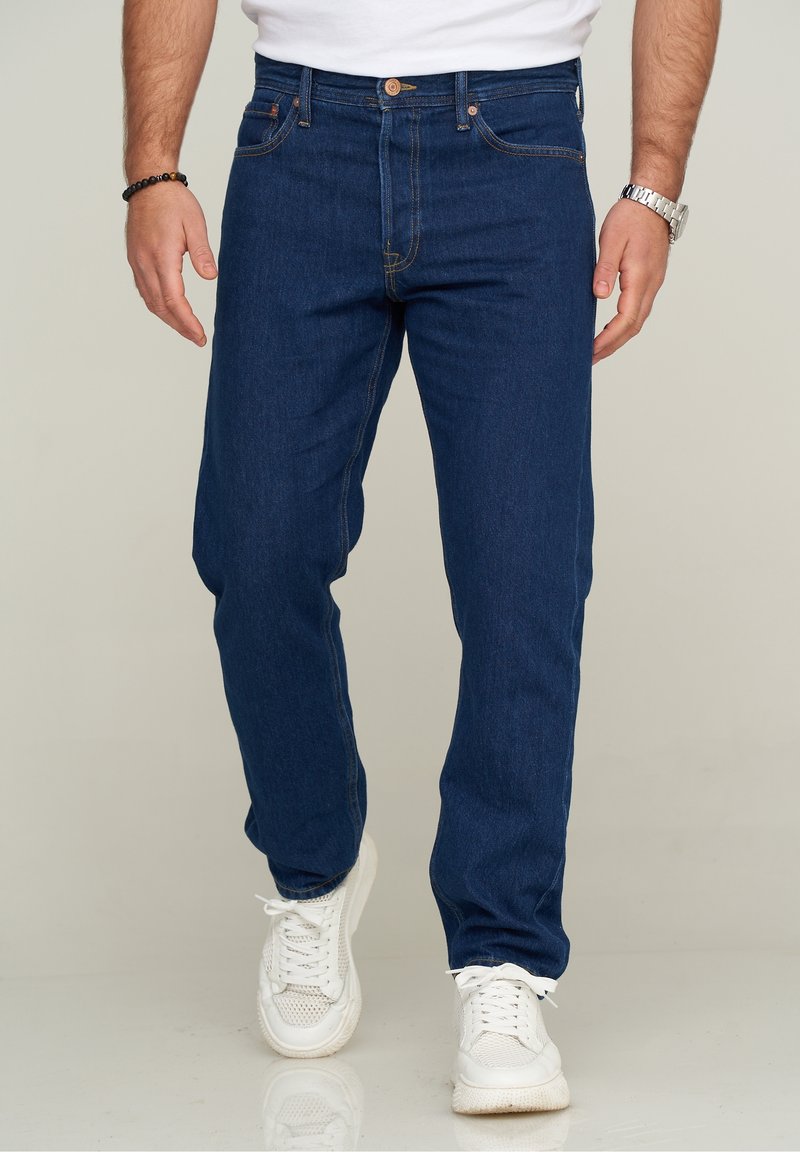 Man som bär mörkblå jeans, vita sneakers, vit skjorta, silverfärgad armbandsklocka och svart pärlarmband står på ett reflekterande golv.