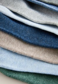 Medžiagų pavyzdžių krūva įvairių spalvų: tamsiai mėlyna, šviesiai mėlyna, smėlinė, pilka ir žalia. Kiekviena medžiaga turi švelnų tekstūrą ir subtilių atspalvių.