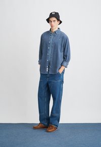 Jeansová košeľa na gombíky, tmavomodrá, v kombinácii s voľnými modrými džínsami. Hnedé topánky a čierny vedierkový klobúk s logom. Jednoduché pozadie.