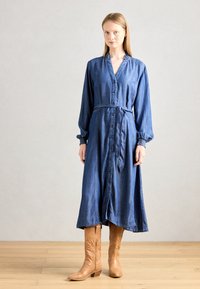 Denim obleka s gumbi in dolgimi rokavi, pasom in tekočo silhueto. Ima globlji izrez in široke manšete. Dopolnjena z rjavimi škornji.