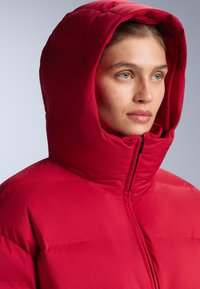 Piros pufi dzseki magas gallérral és nagy kapucnival. Steppelt textúra, látható varrásokkal és elöl cipzáras záródással.